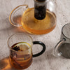Clear glass teapot and mugs with amber tea, garnished with lemon and spices, showcasing elegant designs and color contrast.