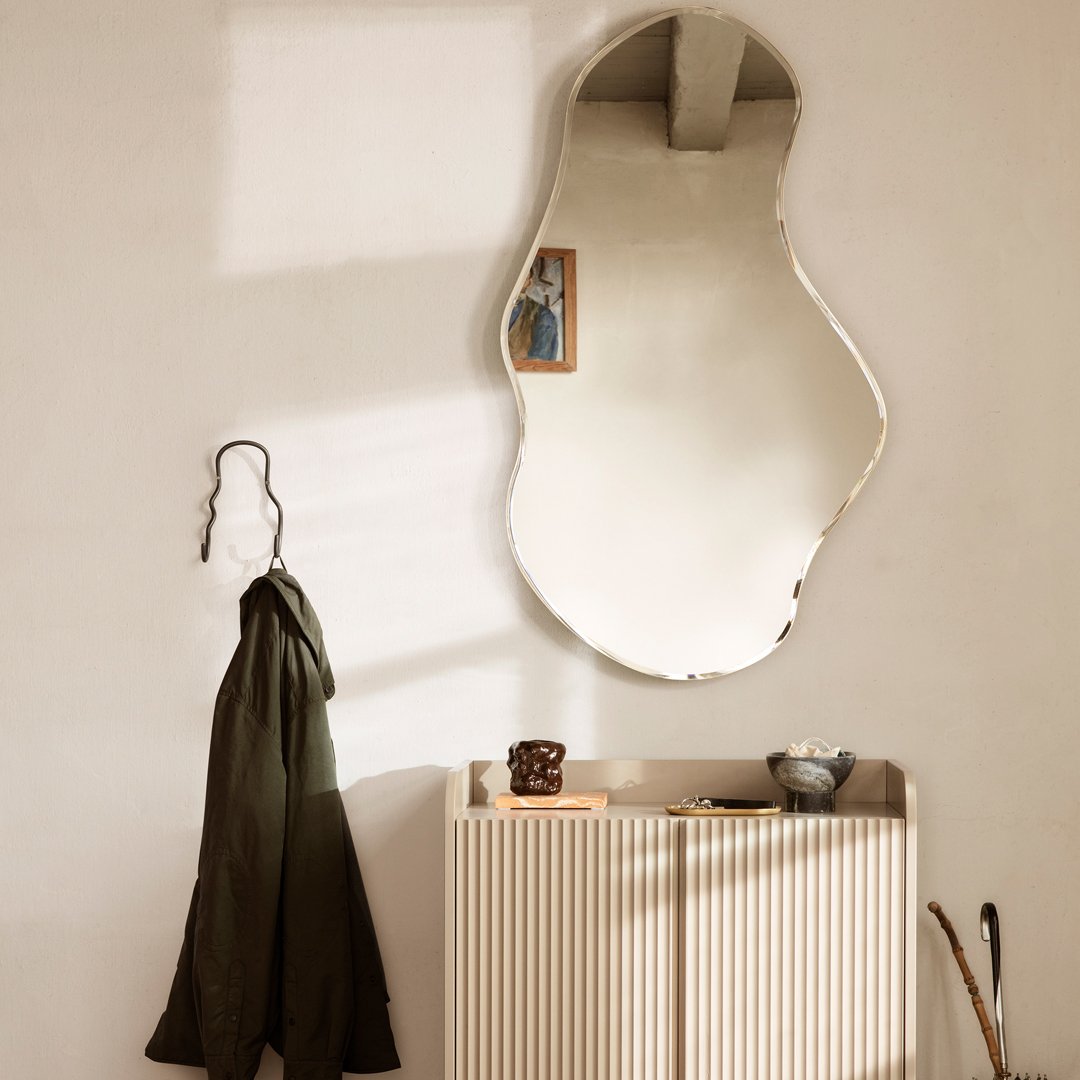 "Unique wavy mirror reflecting soft beige walls with olive green coat and textured cream cabinet, creating a cozy ambiance."