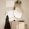 "Unique wavy mirror reflecting soft beige walls with olive green coat and textured cream cabinet, creating a cozy ambiance."