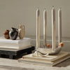 "Modern candlestick holder with four cream candles, surrounded by earthy brown and black pottery on stacked books."