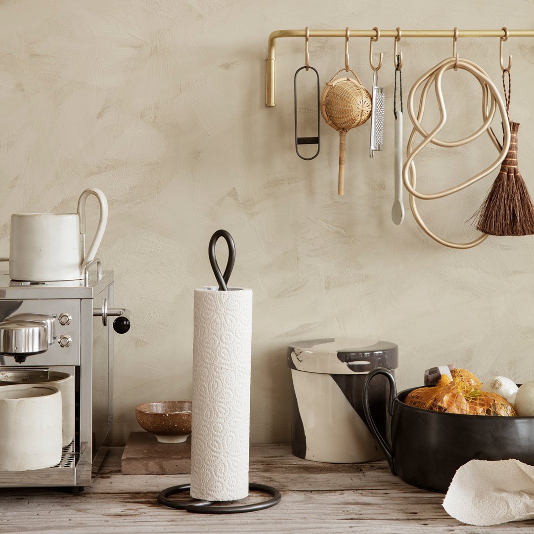 White paper towel holder on a wooden surface, surrounded by neutral tones and various kitchen utensils in muted colors.