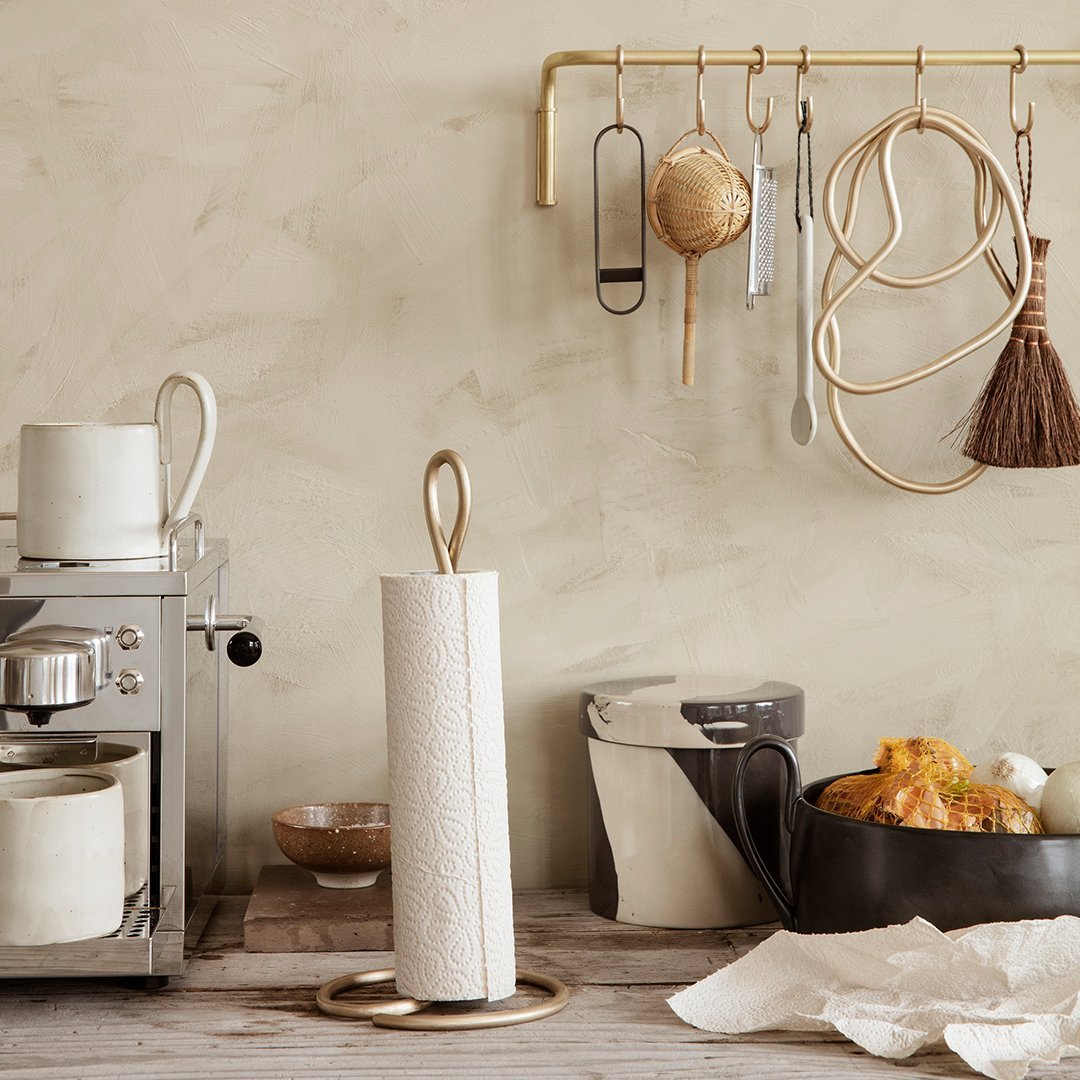 Elegant off-white paper towel holder with intricate pattern against a warm beige backdrop, accented by gold-colored kitchen tools.