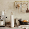 Elegant off-white paper towel holder with intricate pattern against a warm beige backdrop, accented by gold-colored kitchen tools.