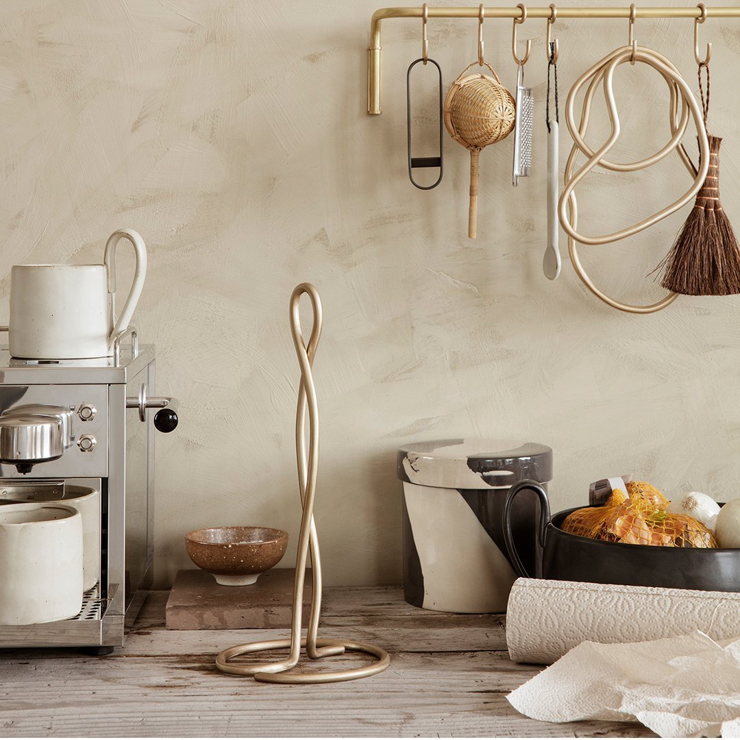 Gold spiral kitchen paper towel holder against a muted beige wall, complementing rustic wooden tones and textures.