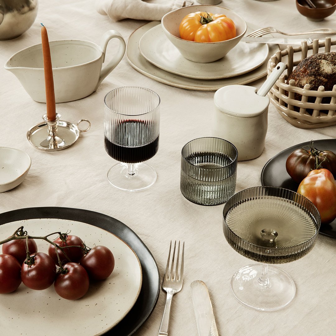 Elegant table setting featuring neutral tones, glassware in smoky hues, and vibrant tomatoes and orange accents.