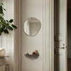 Soft beige walls with a minimalist round mirror and a gray shelf, accented by green foliage and subtle earth tones.