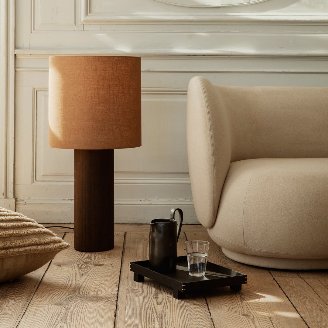 Brown lamp with cylindrical shade, soft beige couch, neutral-toned pillows, wooden floor, minimalistic decor.