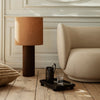 Brown lamp with cylindrical shade, soft beige couch, neutral-toned pillows, wooden floor, minimalistic decor.