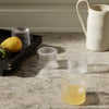 Clear and frosted ribbed glass cups on a marble surface, complemented by a lemon and a simple white pitcher.