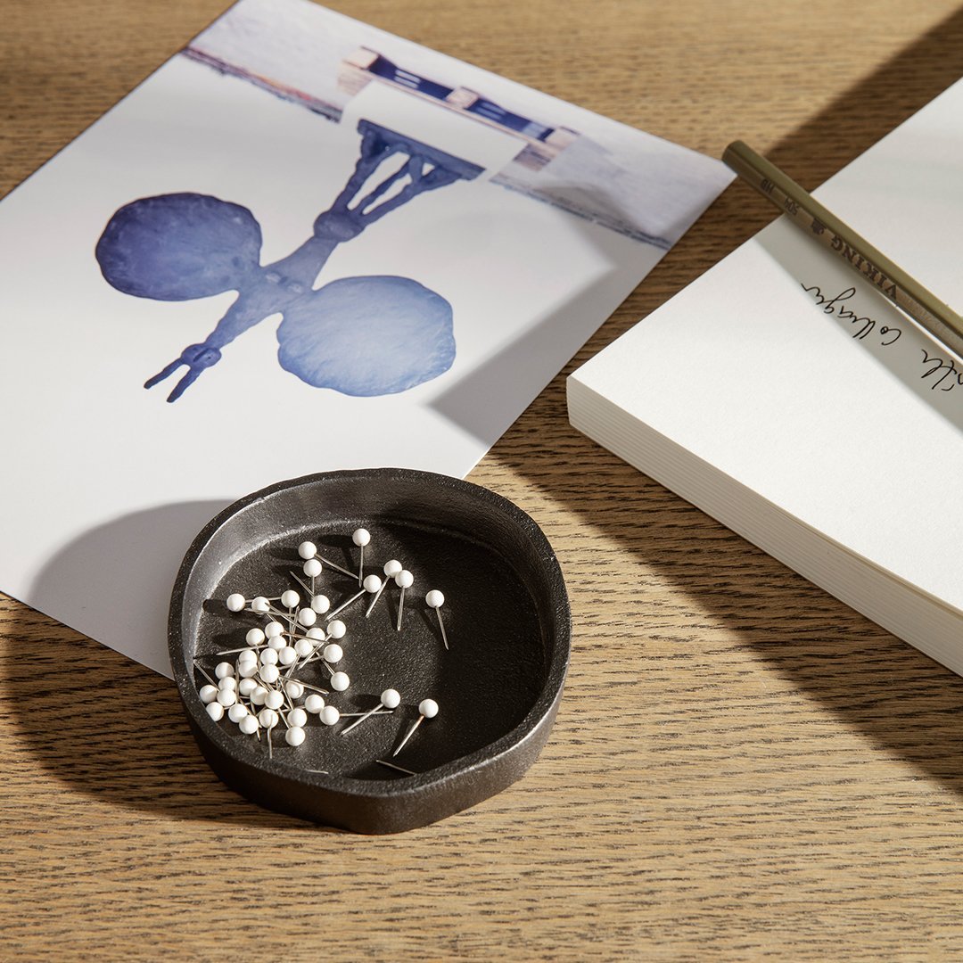 Black round bowl with white pins, set on a wooden surface with soft blue and white artistic prints.