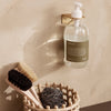 Clear glass soap dispenser with earthy green label, surrounded by natural wooden brushes and a textured woven basket.