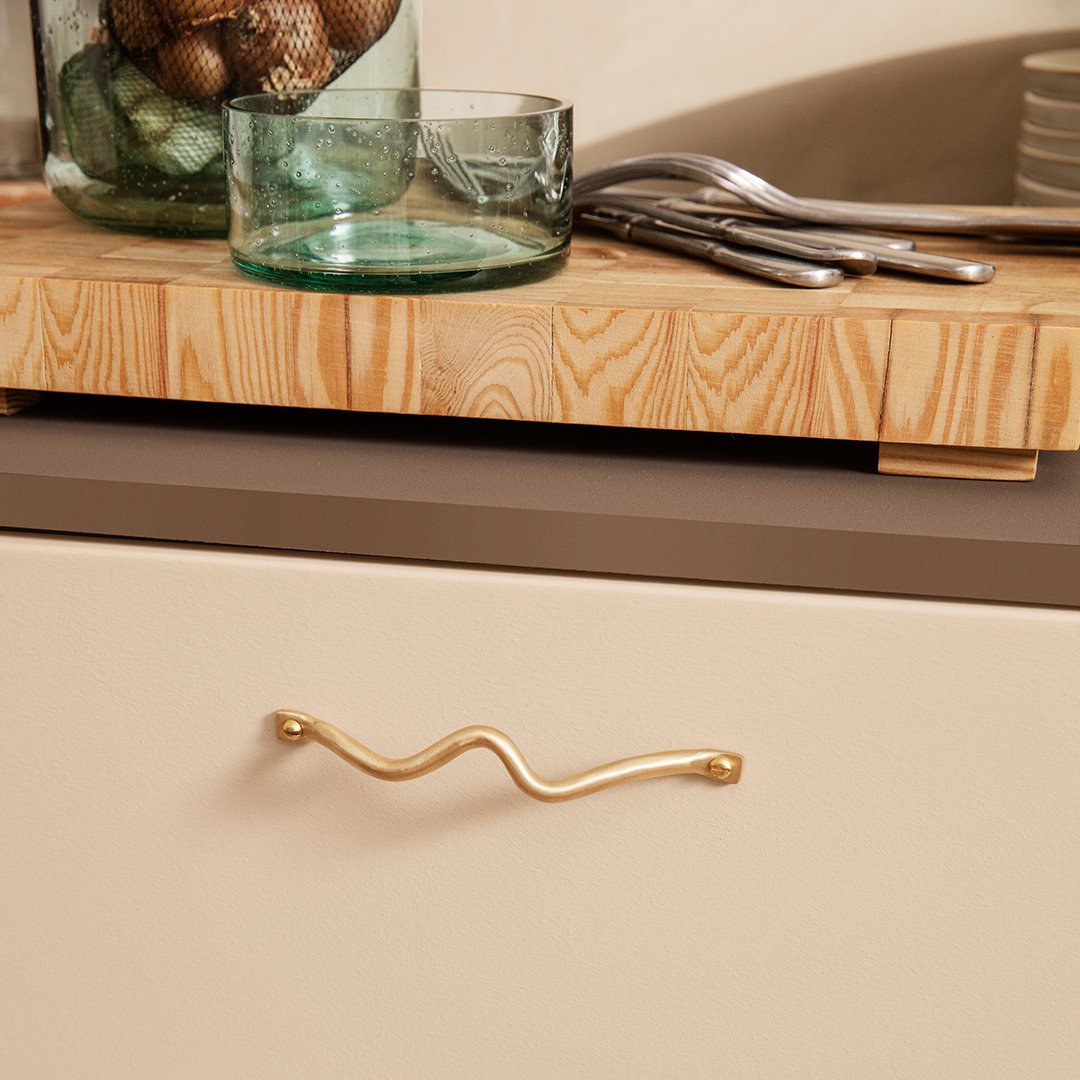 Beige cabinet with wavy gold handle, wooden cutting board, green glassware, and silver utensils on a neutral surface.