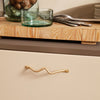 Beige cabinet with wavy gold handle, wooden cutting board, green glassware, and silver utensils on a neutral surface.