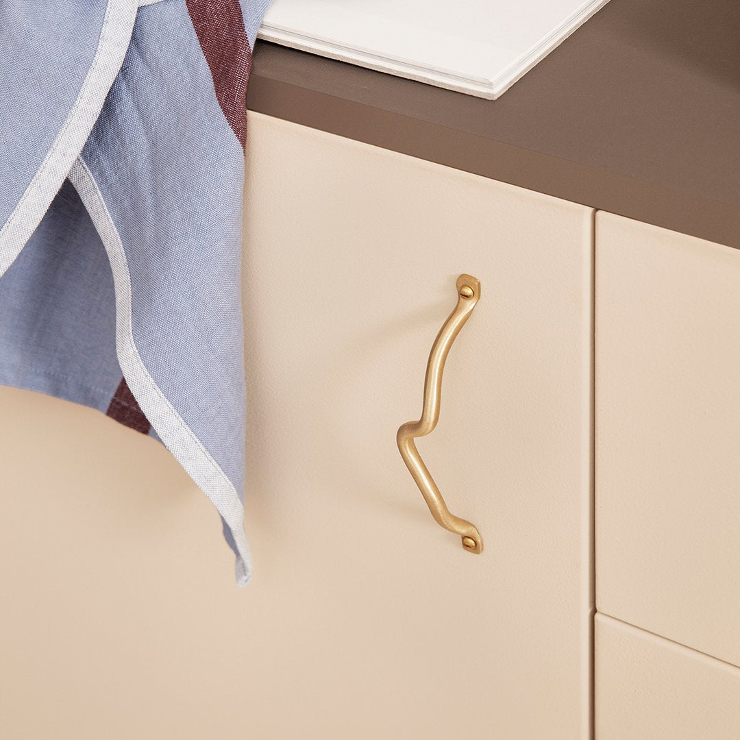 Elegant gold drawer handle on cream cabinet with soft blue and maroon linen towel draped nearby. Modern color palette.
