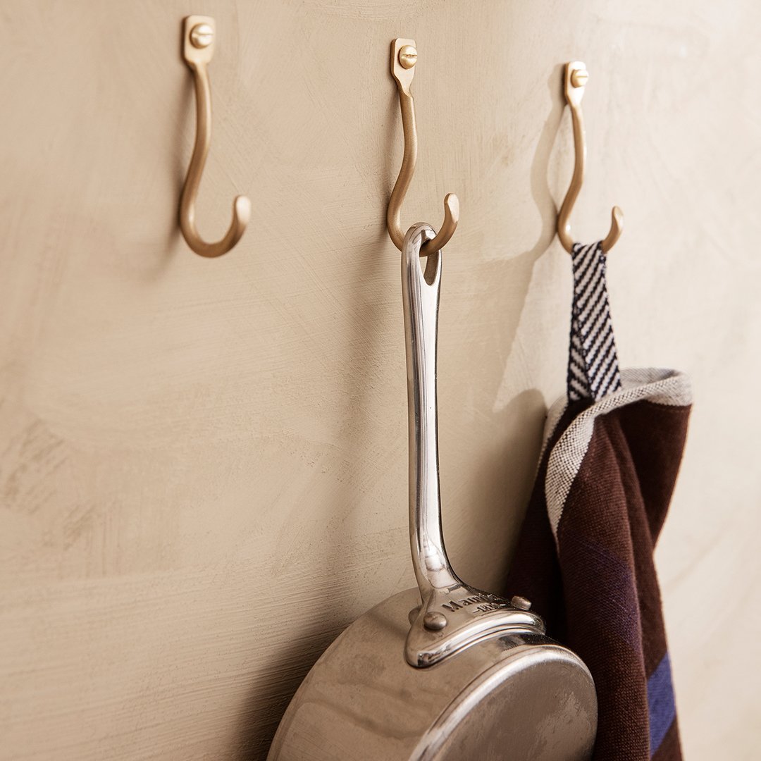 Brushed gold hooks on a beige wall with a silver pot and striped apron in rich brown and white patterns.