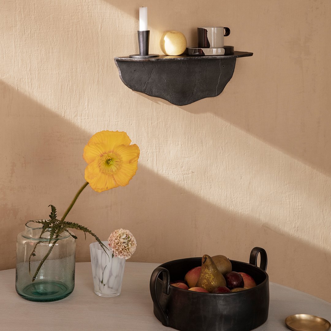 Black wall shelf with natural textures, vibrant yellow flower, earthy tones, and assorted fruits in dark bowl.