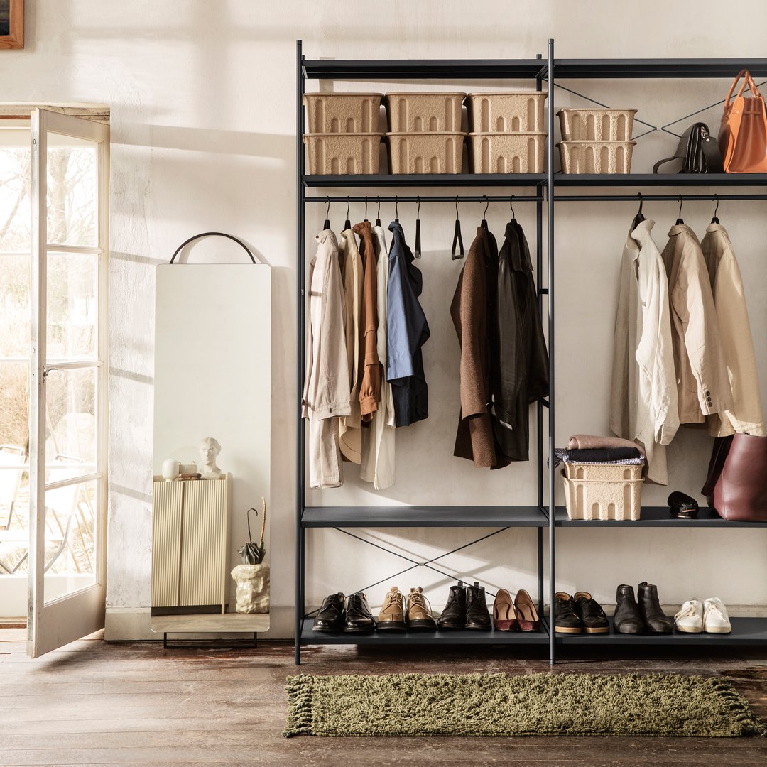 Black metal shelving with beige baskets and assorted clothing. Earthy tones with a green rug and reflective mirror.