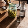 Clear glass jar with green olives, textured gray vases, and earthy tones on a wooden table.