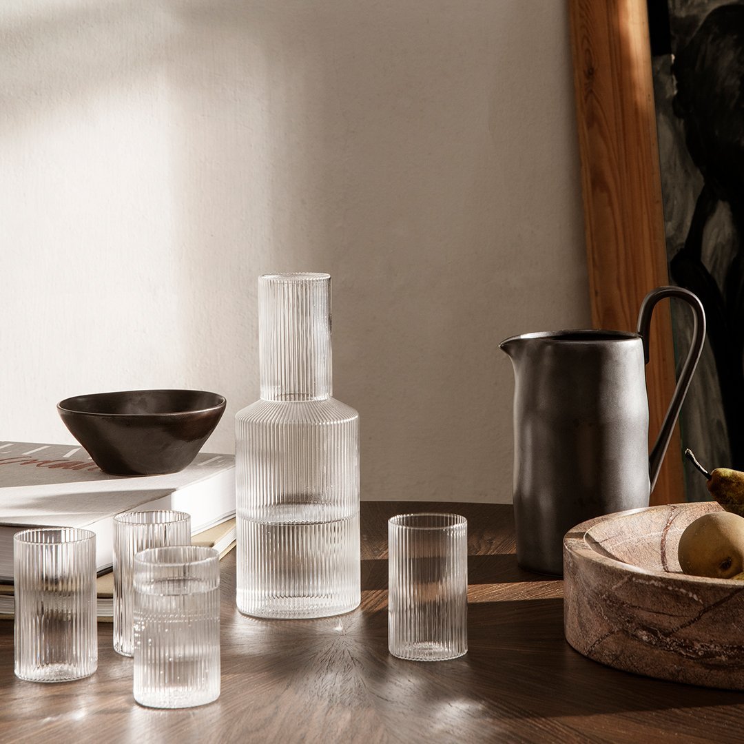 Clear textured glass pitcher and tumblers with a dark ceramic bowl, set on a wooden table. Natural earthy tones.