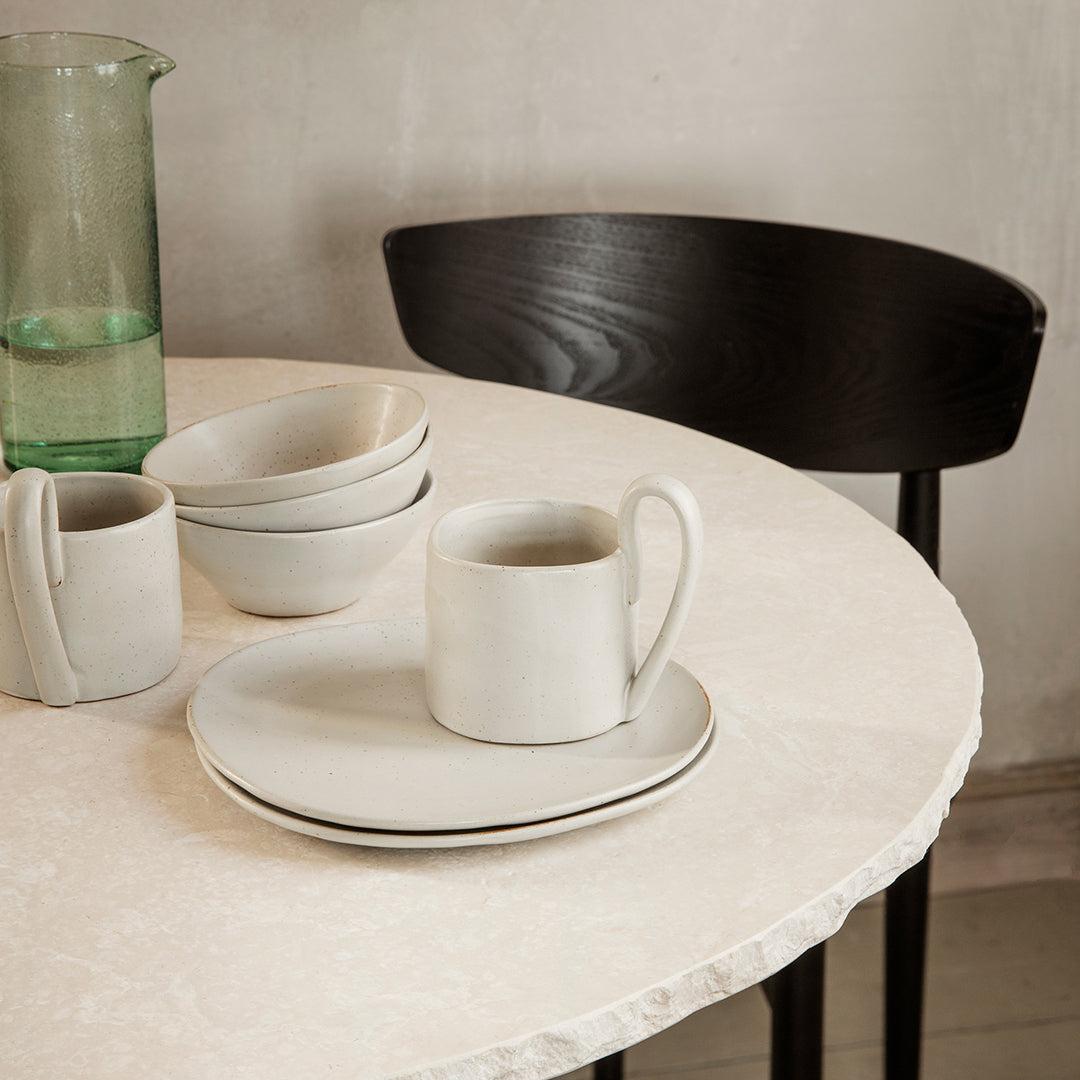 Neutral-toned dishware with subtle black rim, set against a light marble table and wooden chair for a minimalist vibe.
