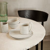 Neutral-toned dishware with subtle black rim, set against a light marble table and wooden chair for a minimalist vibe.