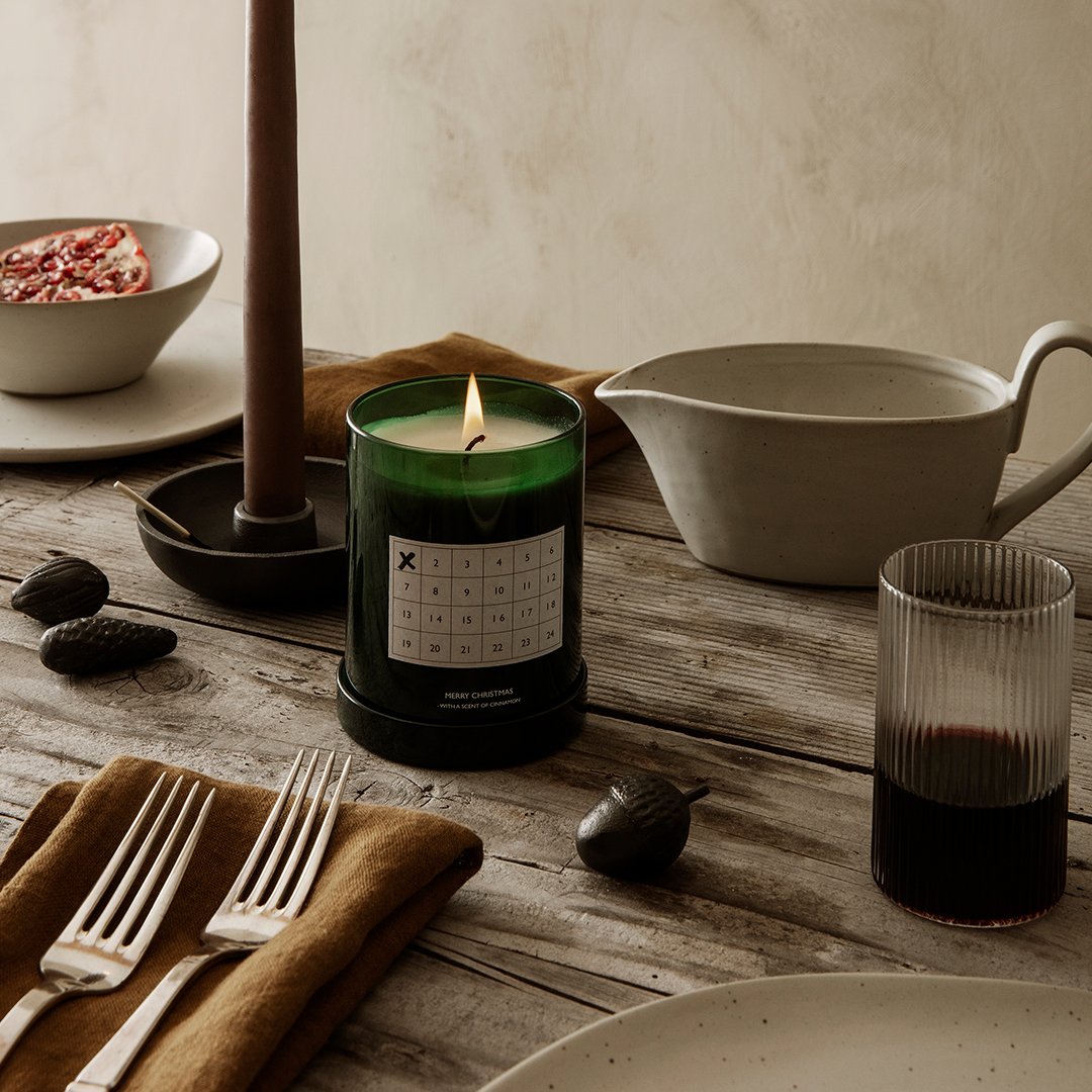 Green glass candle with minimalistic label, set on a rustic wooden table with neutral tones and earthy textures.