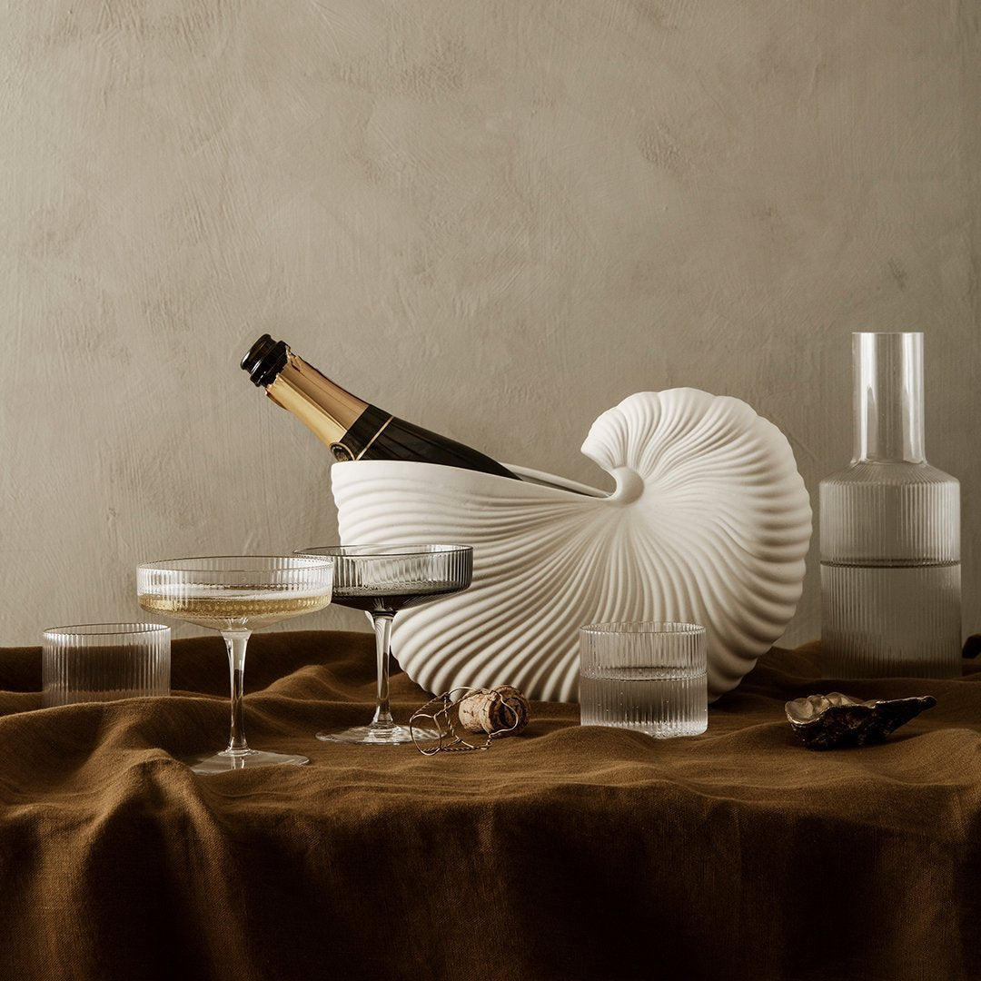 Elegant white shell-shaped wine cooler with rippled texture, accompanied by clear glassware on a rich brown fabric backdrop.