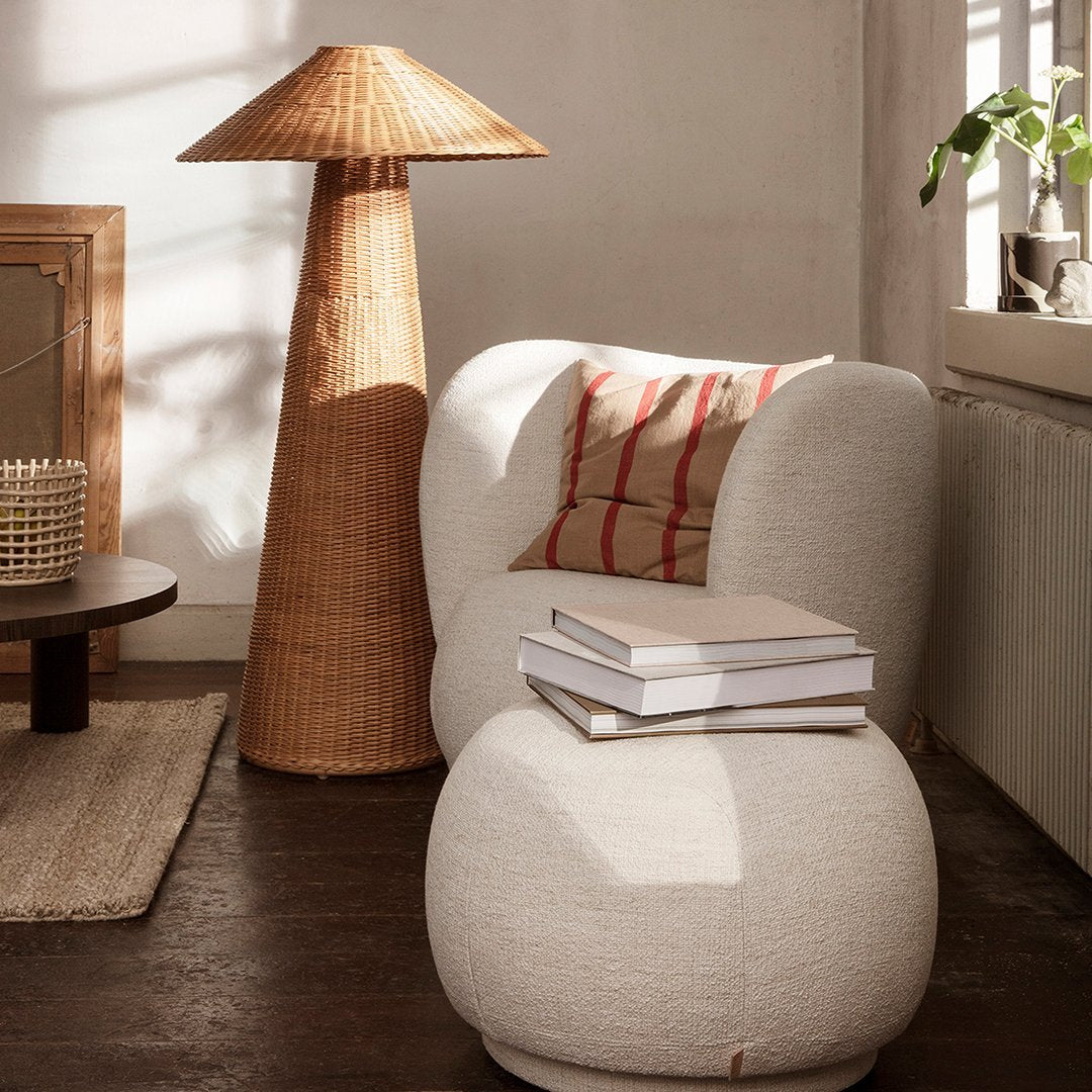 Soft cream upholstered chair with striped red accent pillow, round white ottoman, and natural woven lamp. Cozy neutral tones.