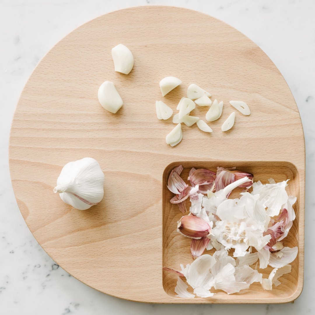 Light wood cutting board with natural grains, featuring white garlic cloves and pinkish garlic skins.