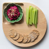 Colorful wooden platter with green bowl, vibrant pink dip, fresh celery sticks, and golden-brown crackers in a playful arrangement.