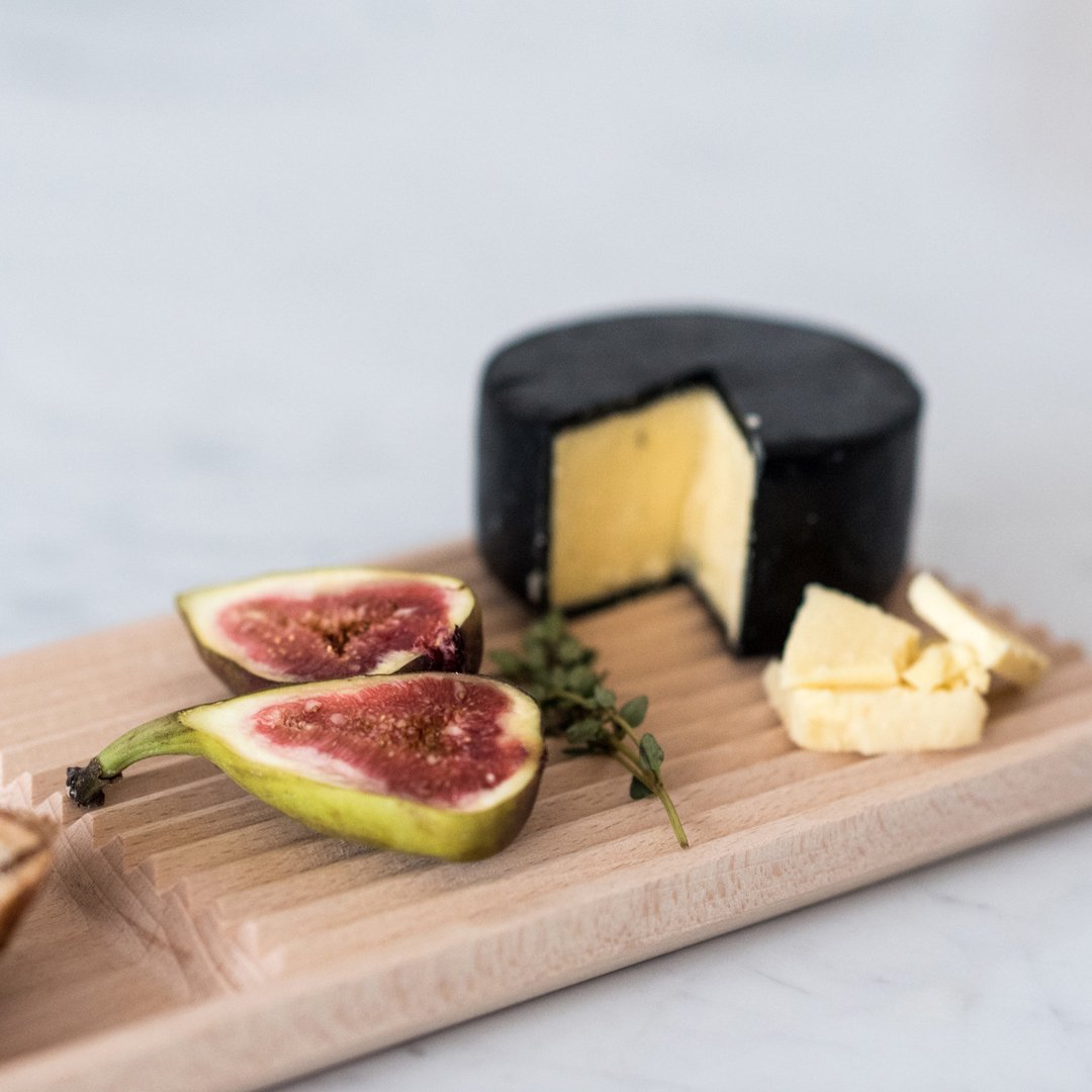 Black cheese wheel, creamy yellow interior, garnished with green figs and brown butter on a wooden board.