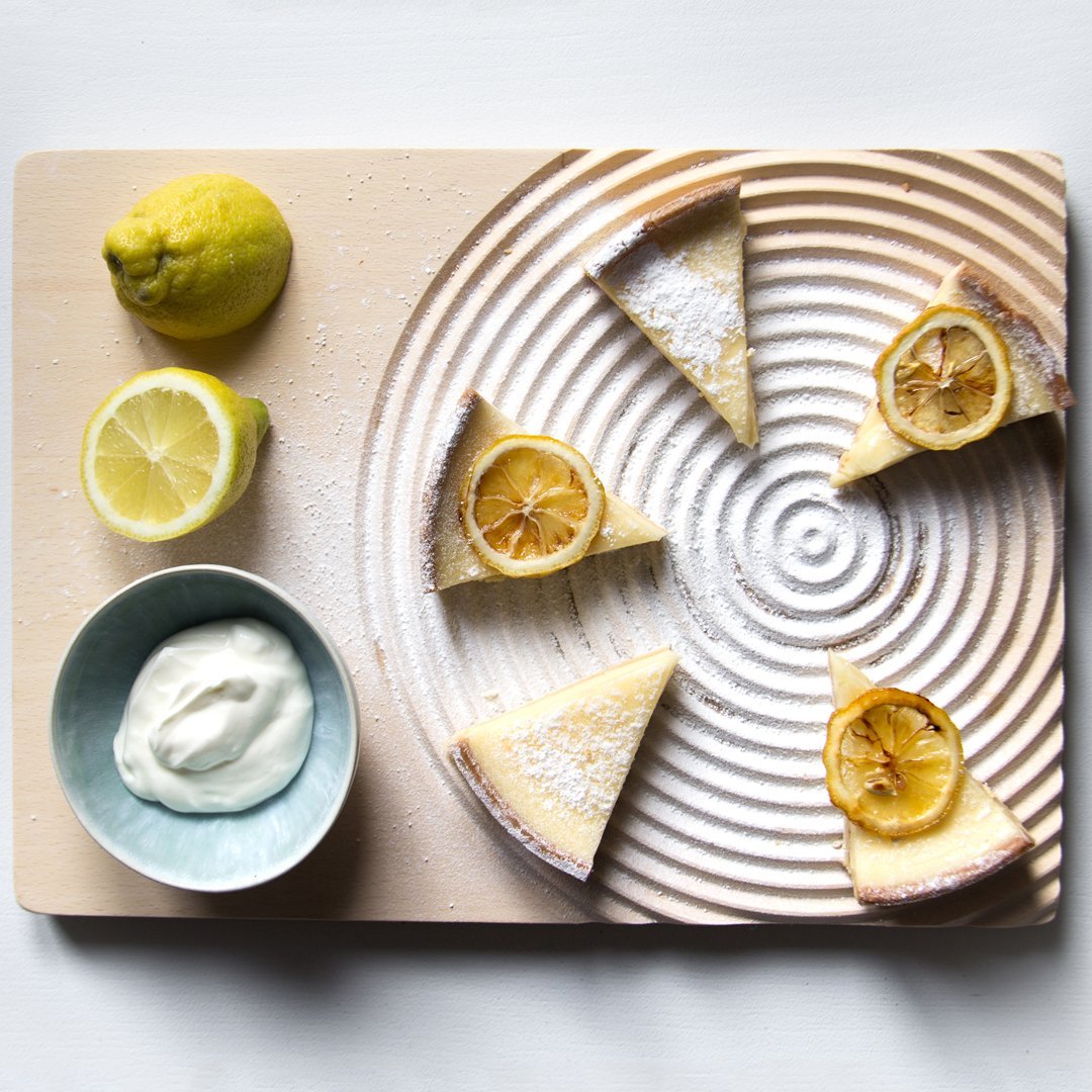Lemon cheesecake slices arranged on a textured cream-colored board, with vibrant yellow lemons and a teal bowl.