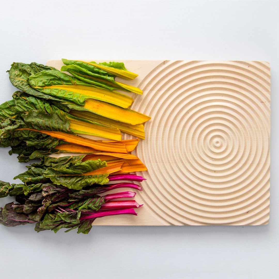 Colorful Swiss chard in yellow, orange, and pink, arranged on a textured wooden cutting board with concentric circles.