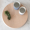 "Beige circular serving tray with spiral pattern, paired with striped blue and white mugs and fresh rosemary."
